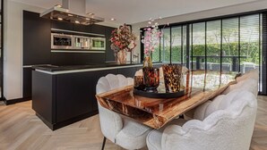 Une cuisine moderne dotée d’un îlot central, d’un coin repas avec une table en bois et des chaises blanches, et de grandes fenêtres offrant une vue sur la verdure.