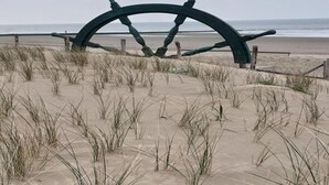 Une grande roue de navire sur une plage de sable, avec de l’herbe au premier plan.