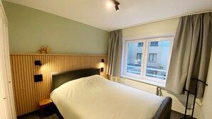 Quarto de hotel com uma cama, um banquinho pequeno, uma janela com cortinas e uma luminária na parede.