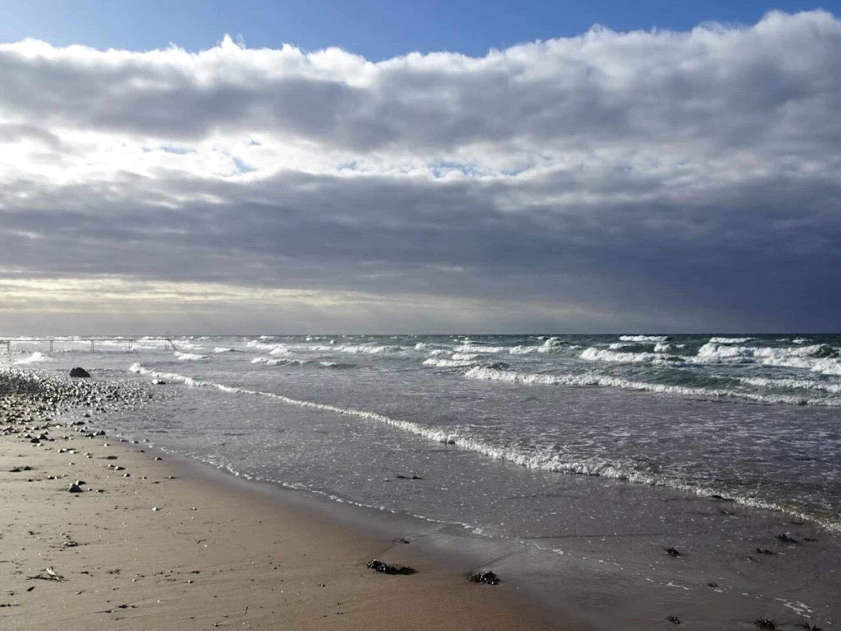 Een strand met golven, een pier in de verte, en een bewolkte lucht.