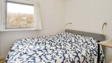 Un dormitorio con cama, una ventana con cortinas, una mesita de noche y una lámpara.