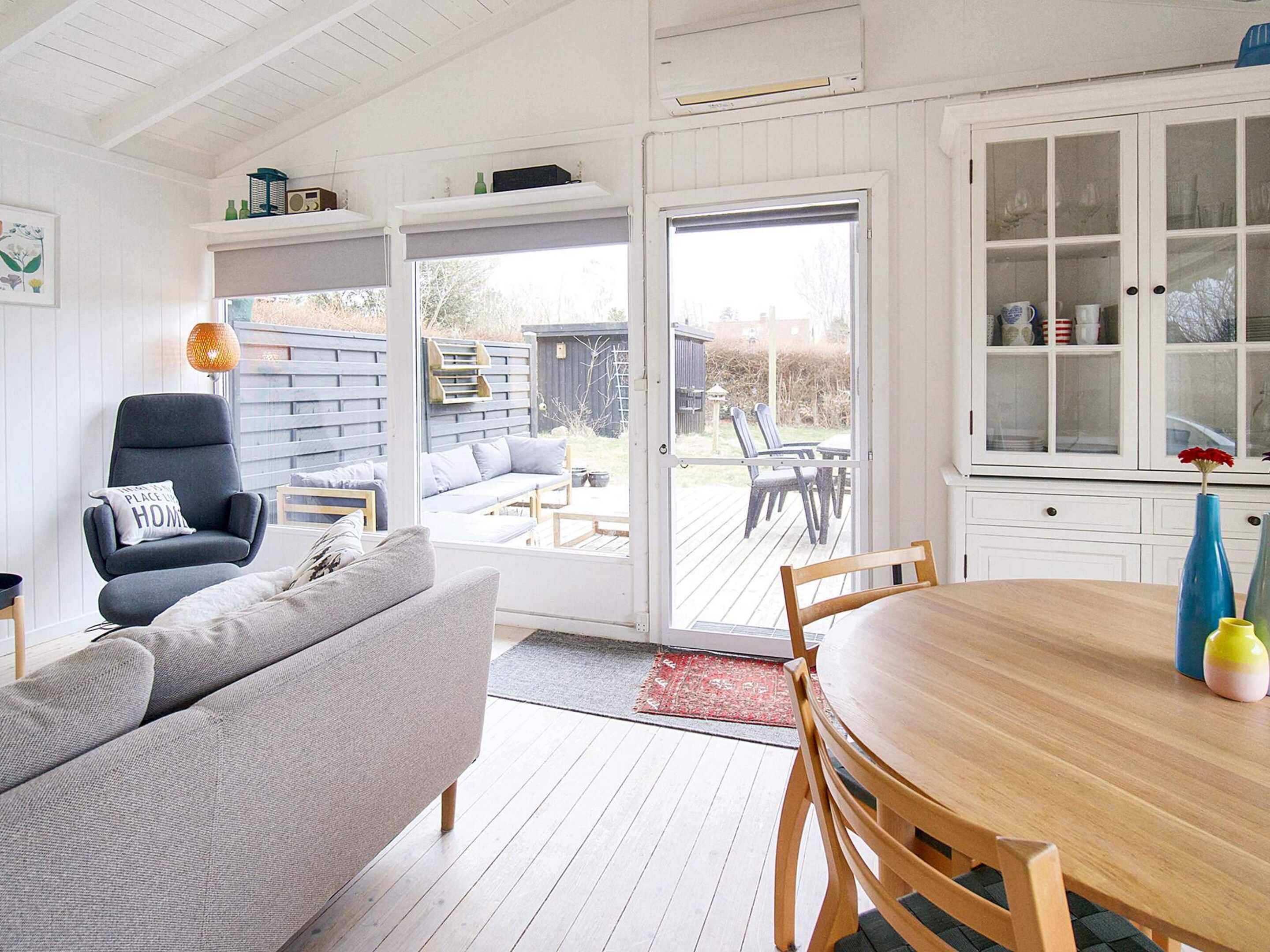 Une pièce comprenant une table à manger en bois, un canapé gris, une chaise et une porte-fenêtre coulissante donnant sur une terrasse extérieure.