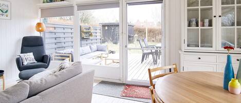 Une pièce comprenant une table à manger en bois, un canapé gris, une chaise et une porte-fenêtre coulissante donnant sur une terrasse extérieure.