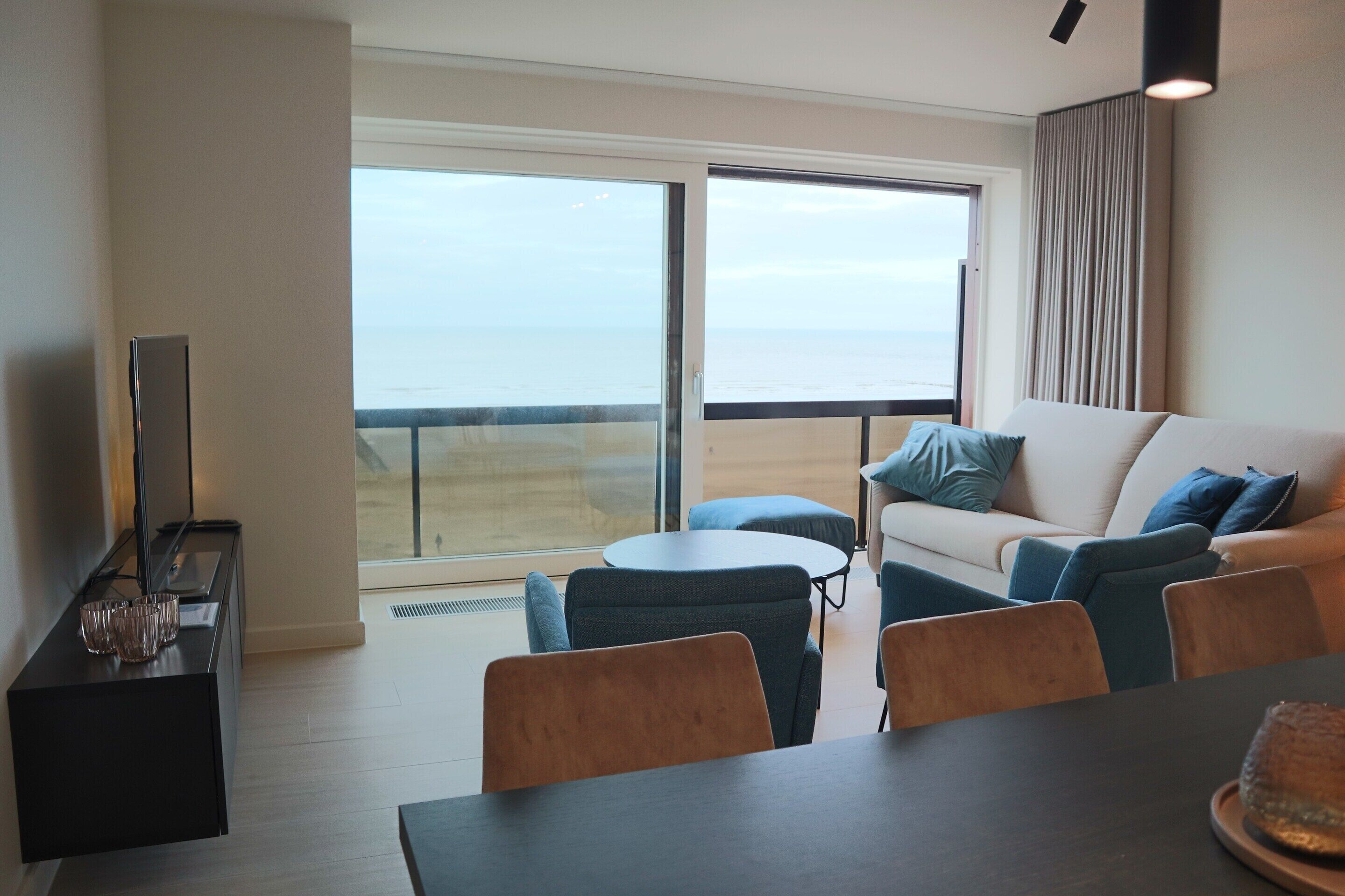 Een moderne woonkamer met een bank, twee fauteuils, een salontafel en een eettafel. Er is een groot raam met uitzicht op zee.