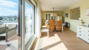 Een ruime woonkamer met een grote schuifpui naar een balkon, een eethoek met rieten stoelen en een witte kast met lades.