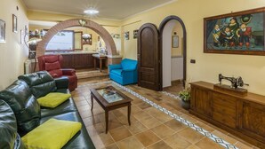 Ein geräumiges Wohnzimmer mit einer Ledersofa, einem blauen Sessel und einem Couchtisch. Es gibt eine Küchenzeile mit einer Bar und einem Esstisch. Der Raum verfügt über einen Fliesenboden und ein großes Gemälde an der Wand.