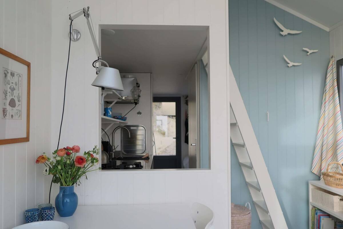 A room with a white table, a blue vase with flowers, a white chair, a bookshelf, and a staircase.