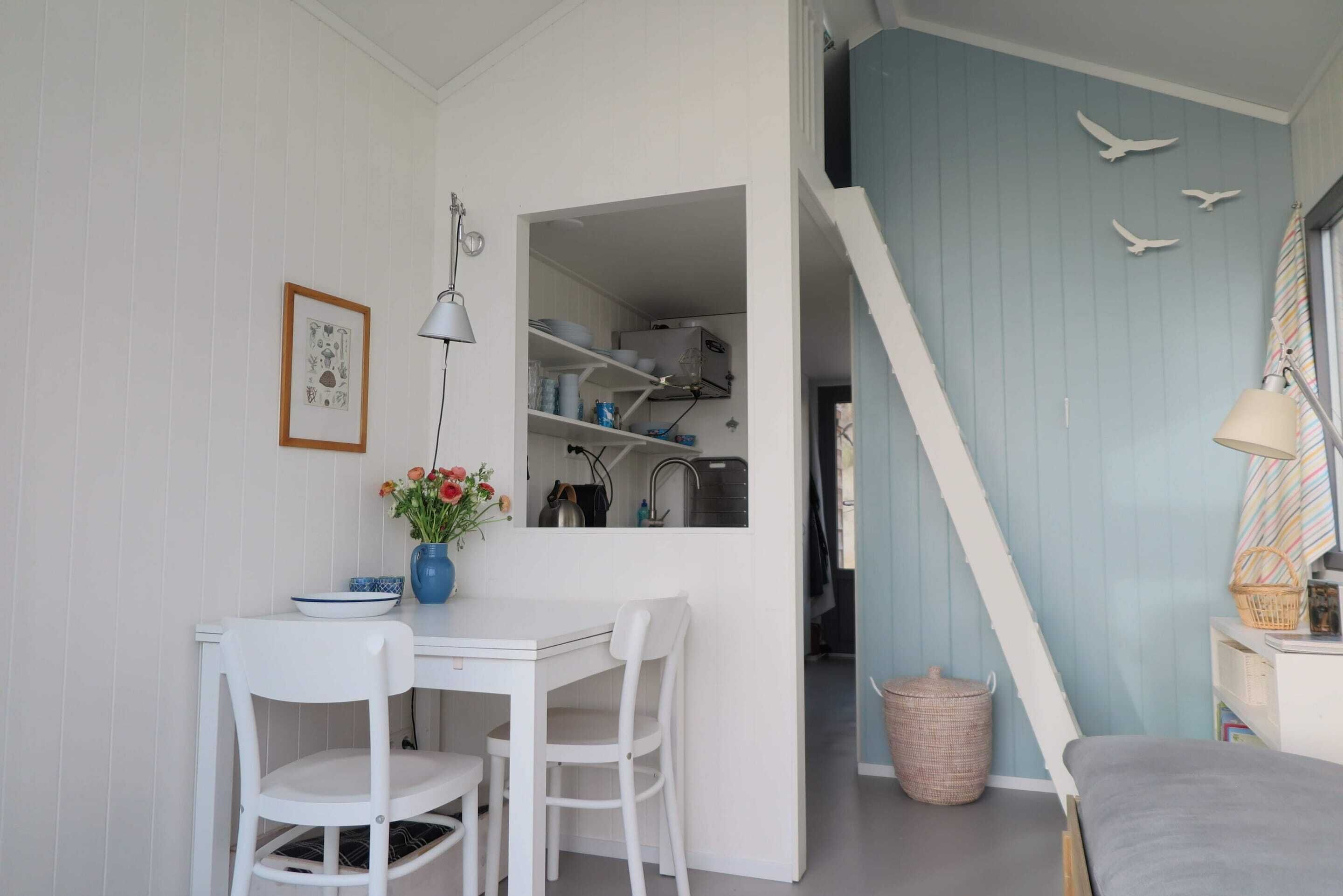 A room with a dining table, chairs, a vase with flowers, a framed picture, and a staircase leading to another room.