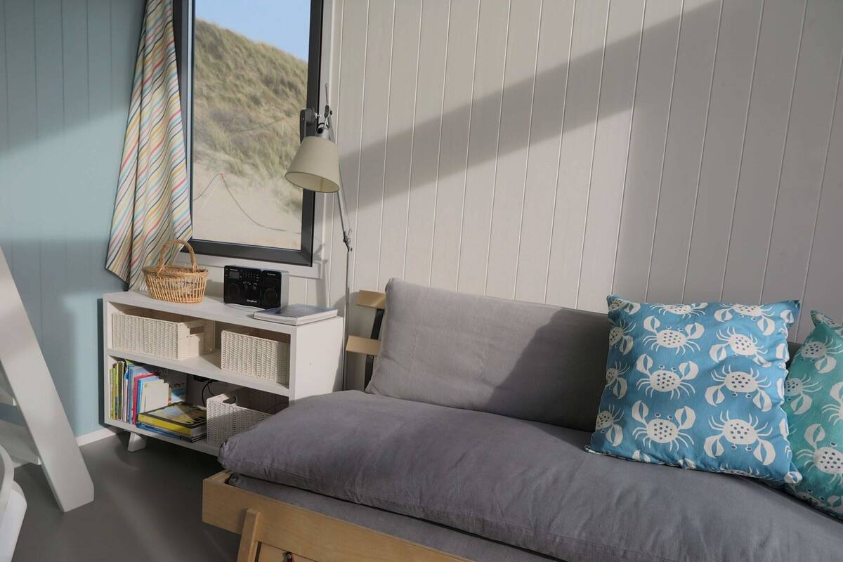 A cozy room with a grey sofa, a wooden side table with a radio, a lamp, and a window with a view of the beach.