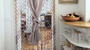 Une porte surmontée d’un rideau noué au centre, qui donne sur une pièce contenant une chaise et une table. On y trouve des étagères remplies de bocaux et un saladier rempli de fruits.