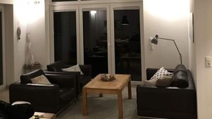 Een woonkamer met een hoge plafond, grote ramen en uitzicht op de keuken. Er is een bank, een salontafel en een lamp.