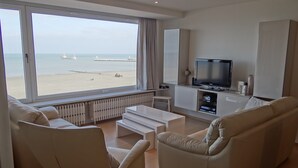 A modern living room with a large window overlooking a beach, a flat-screen TV, and a minimalist design. - Coastal Comfort With Views (Nieuwpoort)