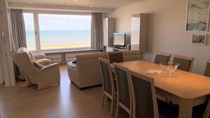 A hotel room with a large window overlooking the beach, a dining area with a table and chairs, and a television on a stand. - Coastal Comfort With Views (Nieuwpoort)