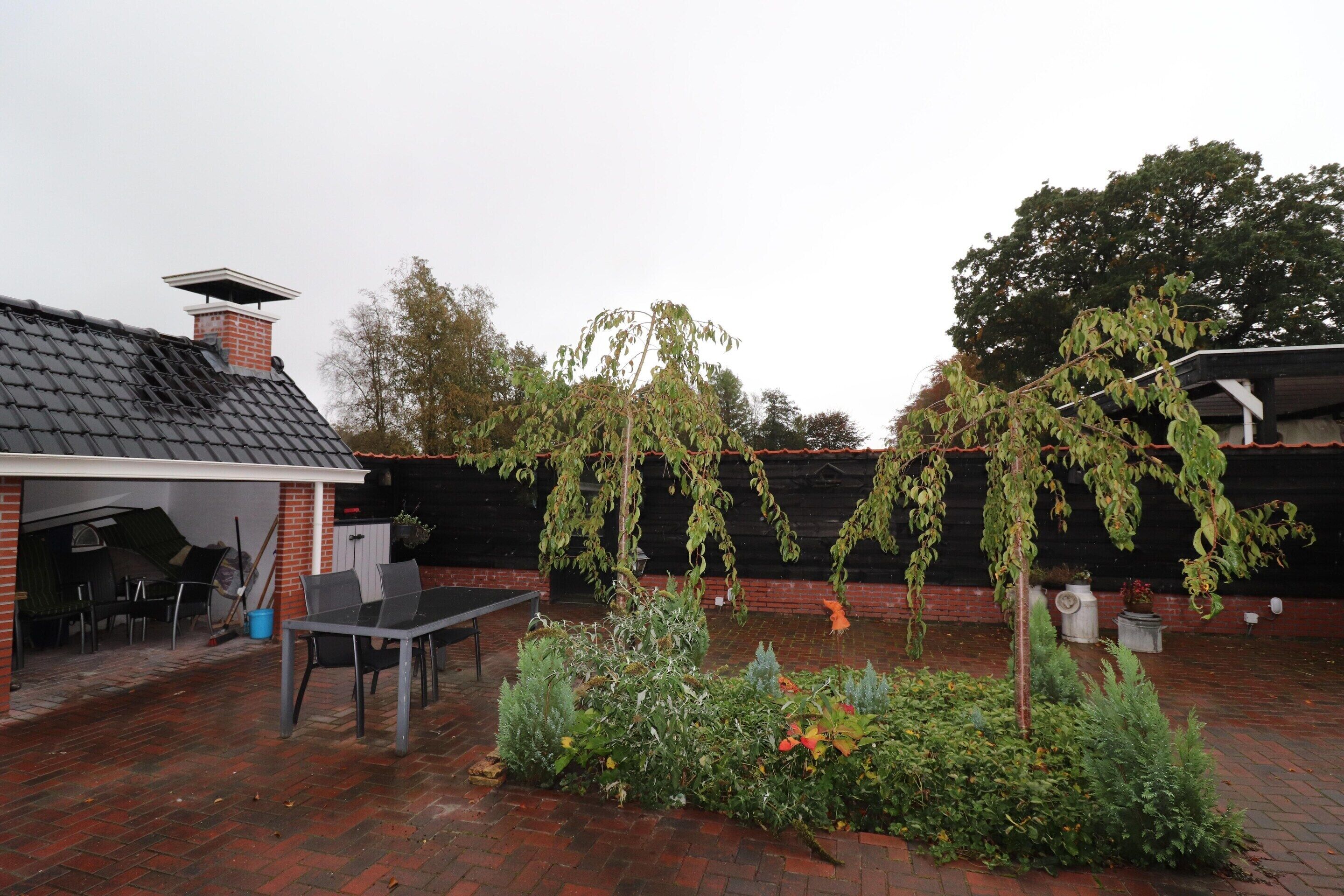 A covered patio area with a brick floor, a table, and chairs. There is a garden with various plants and a tree with hanging branches.