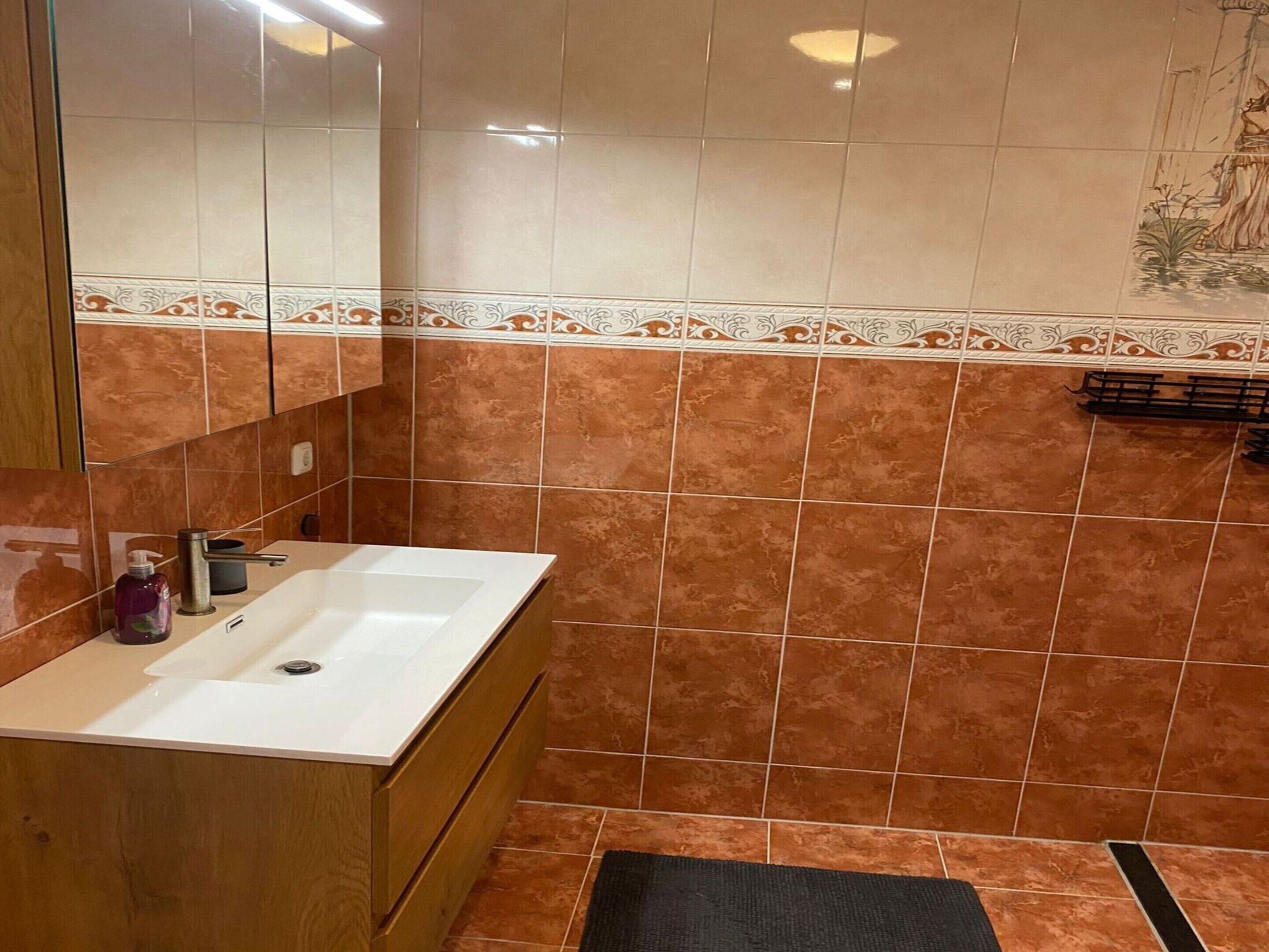A bathroom with a white sink, wooden cabinet, and brown tiled walls.
