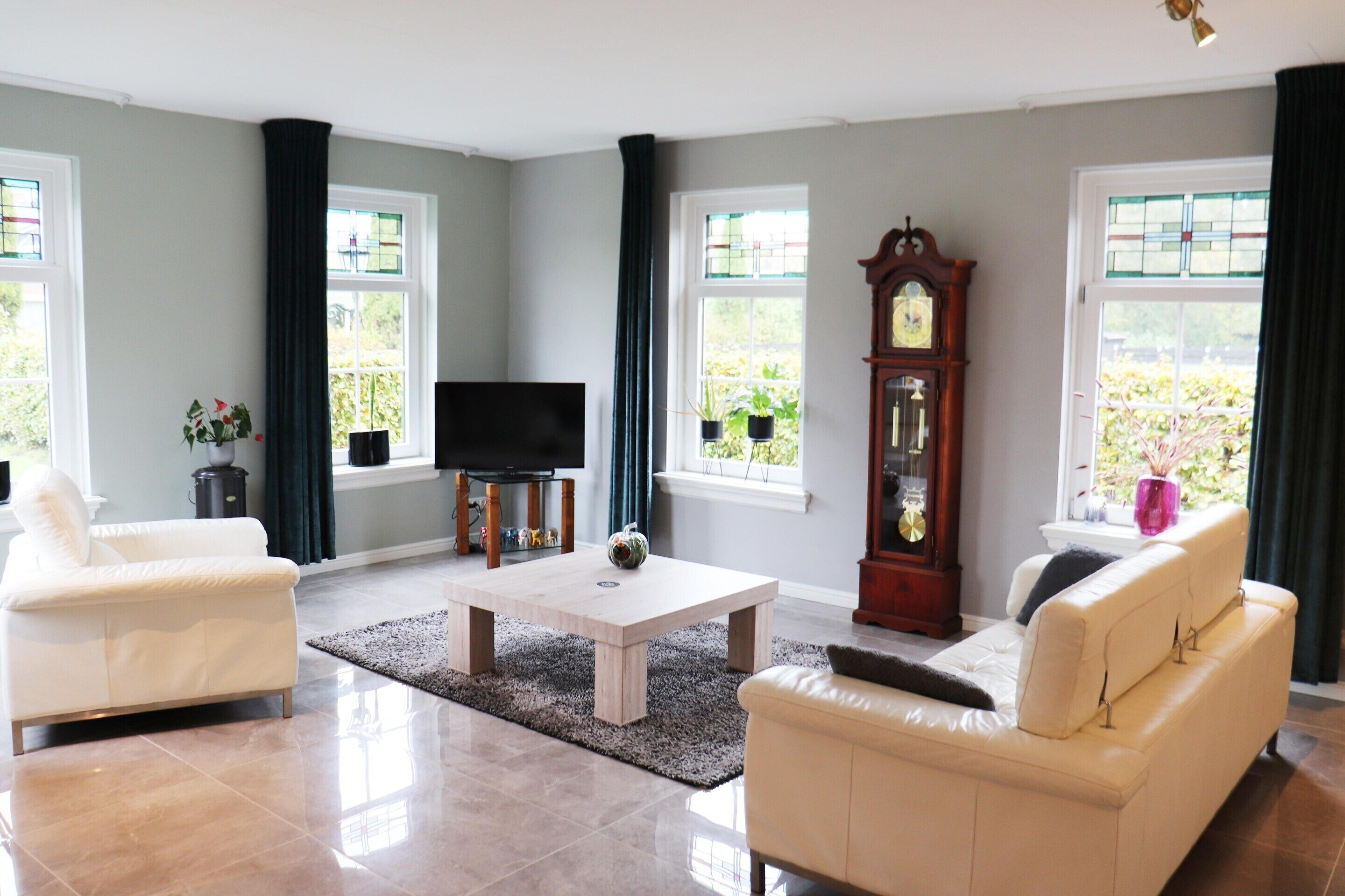 A living room with a white sofa, a coffee table, a TV, a clock, and large windows with curtains.