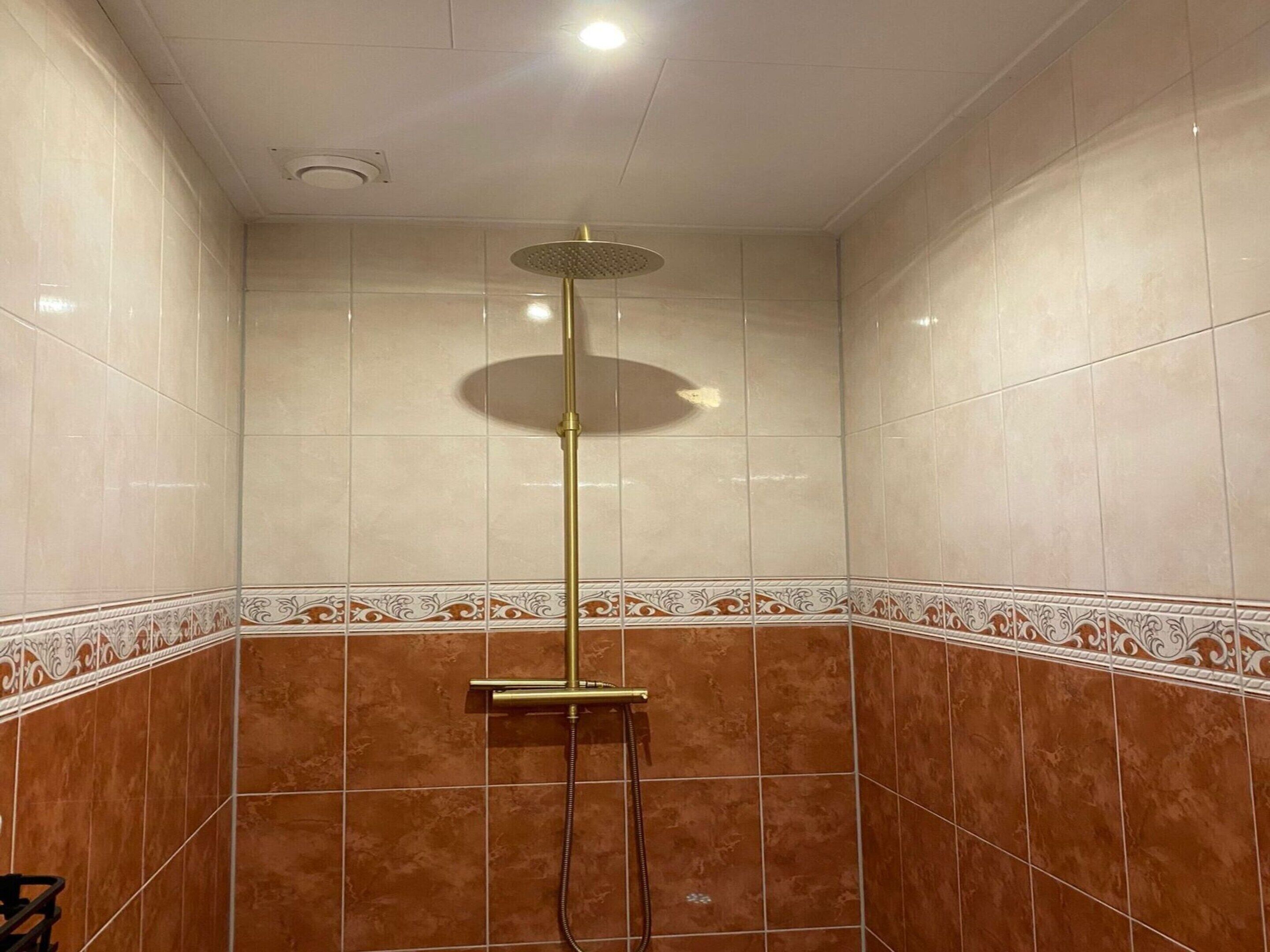 A shower with a gold showerhead and a handheld shower attachment, surrounded by brown and beige tiles with a decorative border.