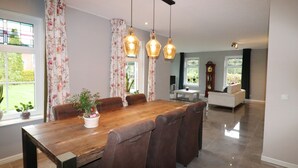 A spacious dining area with a wooden table and brown upholstered chairs, a chandelier, and a view of a garden through large windows. - Apartment in Kornhorn Near Curringherveld (Kornhorn)