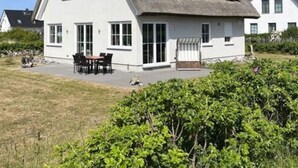 Ein weißes Haus mit grauem Dach, einer Terrasse mit Tisch und Stühlen sowie einem Garten im Vordergrund.