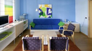 A modern living room with a blue sofa, a coffee table, and two chairs with woven backs. A flat-screen TV and a desk with a lamp are on the left. A colorful abstract painting hangs on the wall. - Sea View and Design Comfortable Vacation Home (Sassnitz)