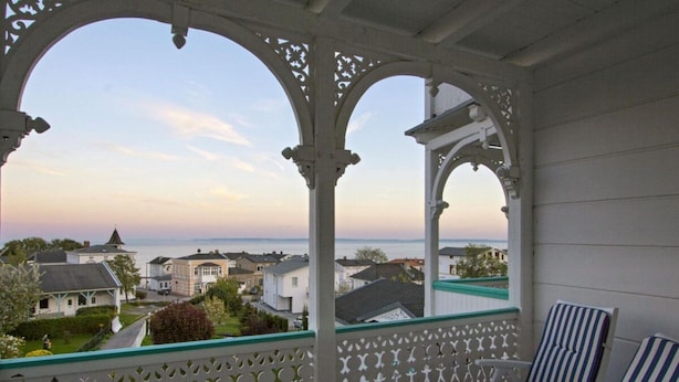 A covered balcony with white lattice railings, blue and white striped lounge chairs, and a view of the sea and buildings in the distance. - Sea View and Design Comfortable Vacation Home (Sassnitz)