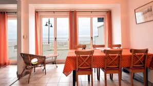 Un comedor con mesa y sillas, una silla de mimbre, vista a la playa y un balcón con barandilla.
