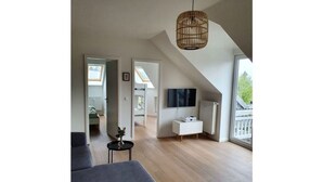 A modern living room with a sofa, a small table, a TV mounted on the wall, and a large window providing natural light.