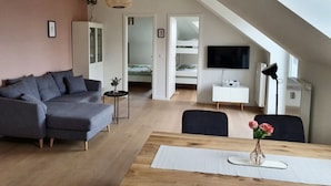 A modern loft apartment with a sloped ceiling, a grey sectional sofa, a dining table with a vase of flowers, a TV mounted on the wall, and a staircase leading to a loft bed area.