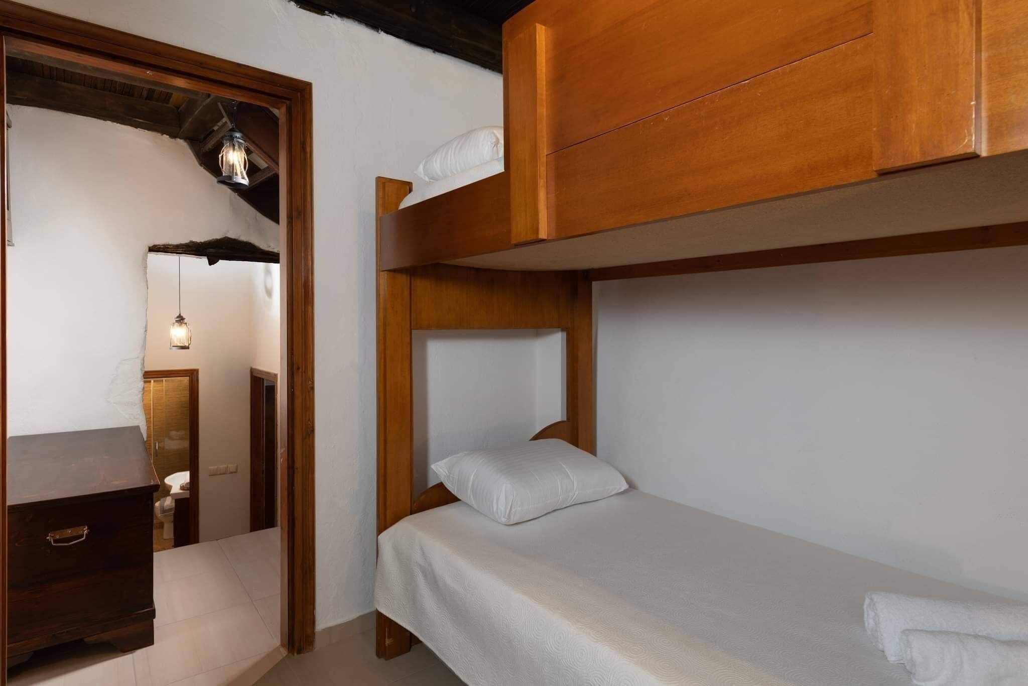 A bunk bed with a wooden headboard and a white mattress. There is a wooden dresser to the left and a doorway leading to a bathroom.