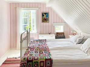 Une chambre avec un plafond en pente, un lit recouvert de draps blancs, une couverture colorée, une table de chevet, une lampe et une fenêtre avec des rideaux.