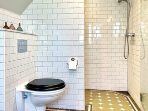 Une salle de bain avec des carreaux de céramique blancs rectangulaires, des toilettes avec un abattant noir, une douche avec une porte en verre et un carrelage au sol à motifs.