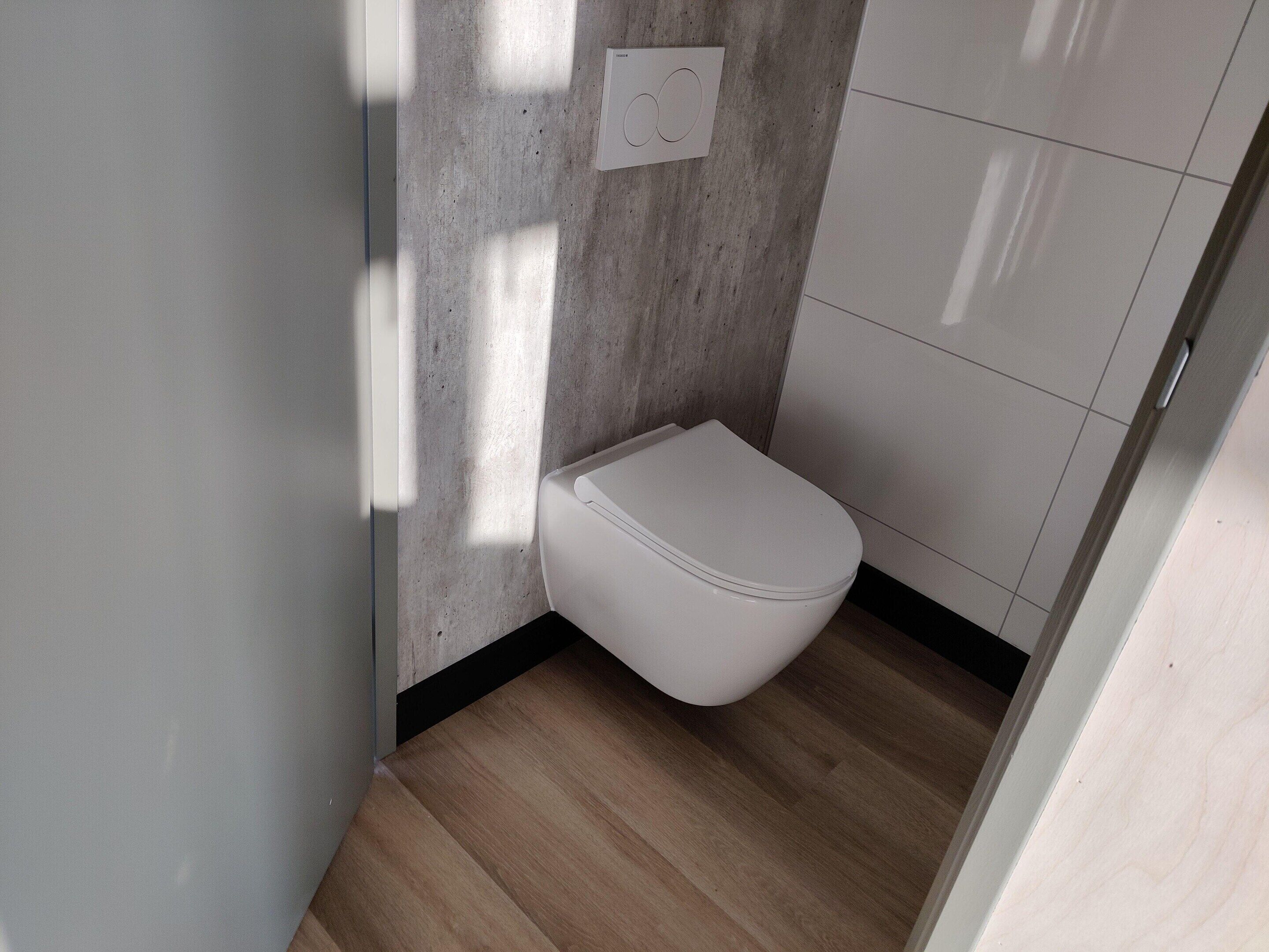 Et moderne badeværelse med et hvidt toilet, lyse trægulve og en væg med betonudsmykning.