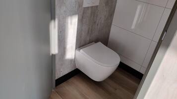 Et moderne badeværelse med et hvidt toilet, lyse trægulve og en væg med betonudsmykning.