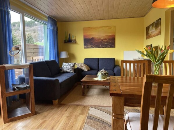 A cozy living room with a sofa, a coffee table, and a dining table with chairs. There is a painting on the wall and a window with curtains. - Holiday Home Lake View (Unterwellenborn)