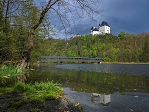 A castle on a hill overlooking a calm lake with a wooden bridge crossing the water.