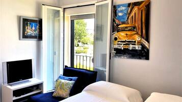 A hotel room with a bed, a TV on a stand, a painting of a train, and a balcony with a view of greenery.