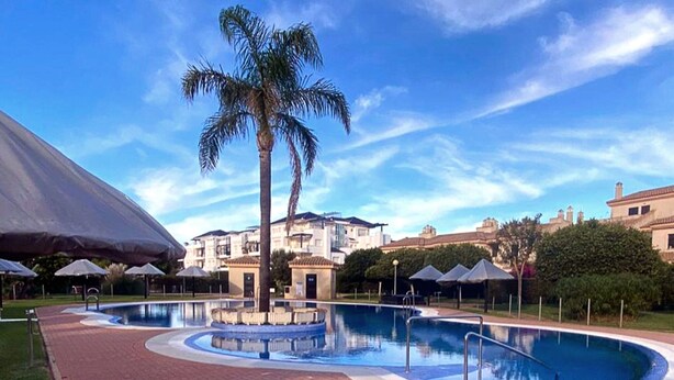 A swimming pool with a palm tree, surrounded by a brick walkway, and buildings in the background. - Belvilla by OYO Holiday Home in Rota (Rota)
