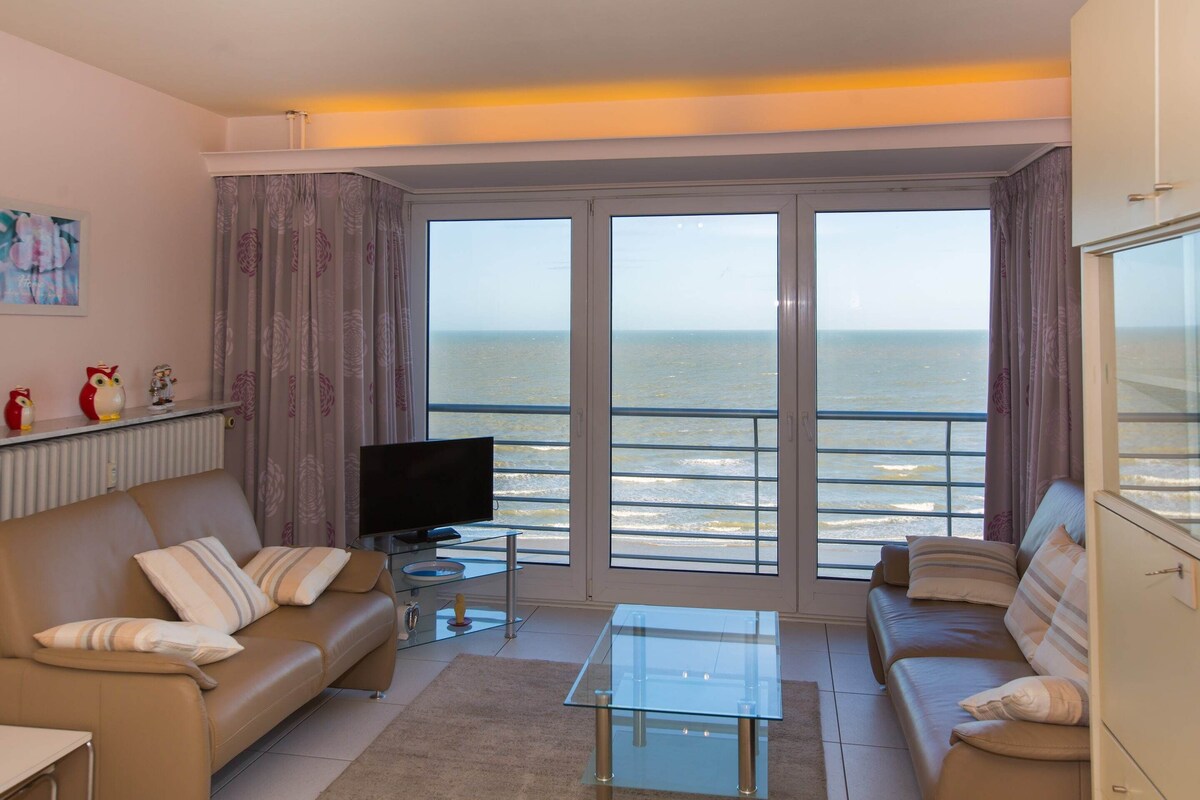 Uma sala de estar com sofá, mesa de centro de vidro e televisão. Há uma janela grande com vista para o mar.