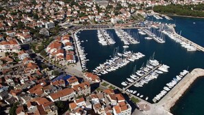 Vista aérea de un pueblo costero con un puerto deportivo lleno de barcos, rodeado de edificios compactos con tejados de tejas rojas.