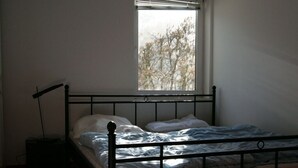 Un lit simple avec un cadre en métal et du linge de lit blanc, placé sous une fenêtre munie de stores, dans une pièce aux murs de couleur unie.