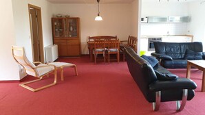 Un salon avec une moquette rouge, des meubles en bois, un canapé noir et un coin repas comprenant des chaises et une table en bois.