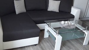 Ein modernes Wohnzimmer mit einem schwarzen Ecksofa, einem Glastisch und einer Küche im Hintergrund.