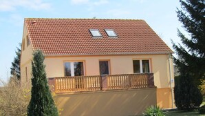 Ein zweigeschossiges Haus mit rotem Ziegeldach, einem Balkon mit Holzgeländer und einem umzäunten Garten.