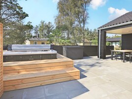Un jacuzzi con escalones de madera que lo conducen, un patio cubierto con mesa y sillas, y vistas a árboles y casas circundantes.