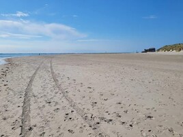 A wide sandy beach with footprints and tire tracks, a clear sky, and a distant building on the right.