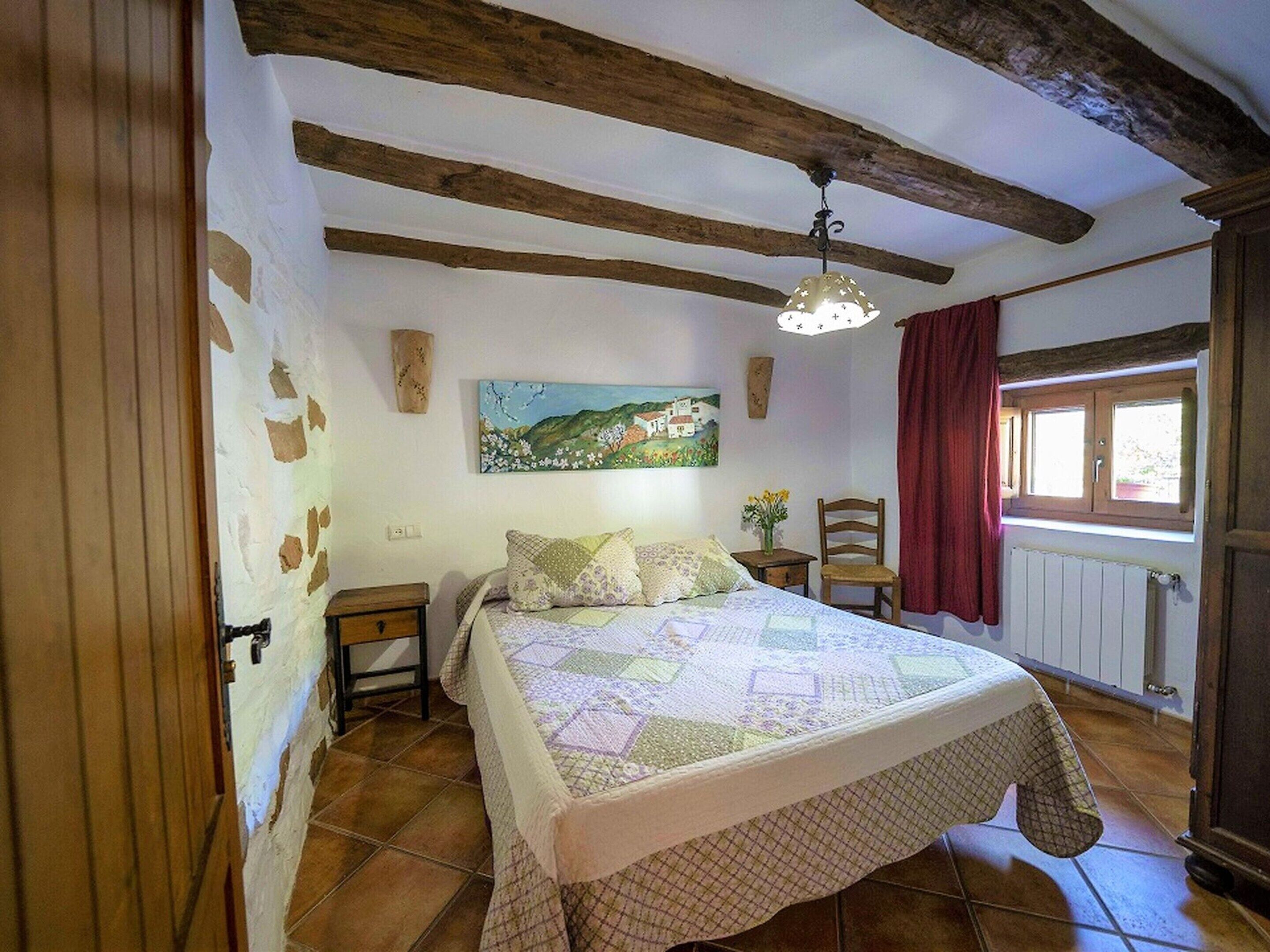 Ein Schlafzimmer mit Holzboden, einem Bett mit Bettdecke, einem Nachttisch, einem Sessel, einem Fenster mit Vorhängen und einem Bild an der Wand.