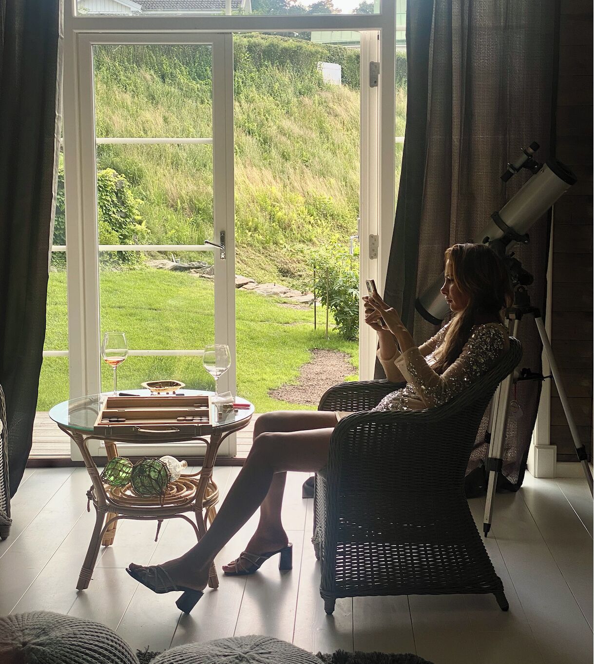 A woman seated by a large window with a telescope, a glass table with a wine glass, and a wicker chair.