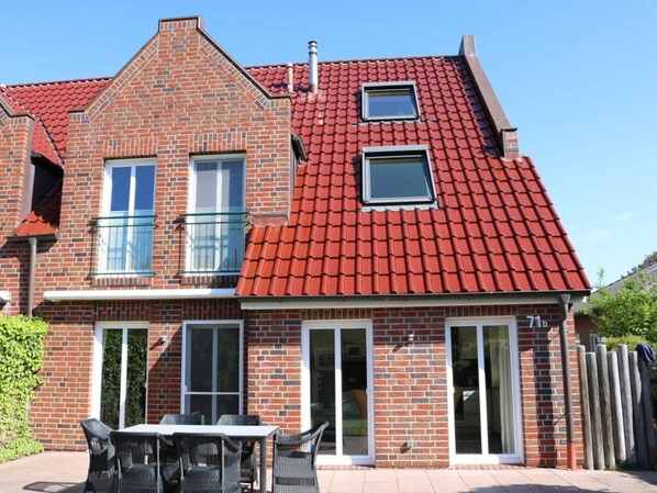Ein zweistöckiges Backsteinhäuschen mit rotem Ziegeldach, einem Balkon und einer Terrasse mit Gartenmöbeln.