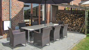 Een terras buiten met een tafel, stoelen en een grote parasol, met een bakstenen gebouw op de achtergrond.