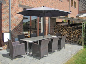 Een terras buiten met een tafel, stoelen en een grote parasol, met een bakstenen gebouw op de achtergrond.